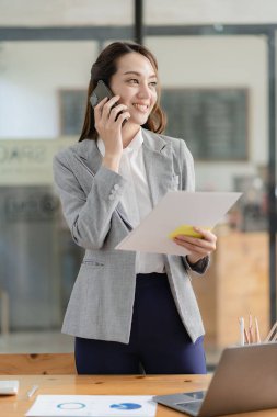 Gülümseyen Asyalı iş kadını masasının önünde akıllı telefonuyla iş tartışıyor.