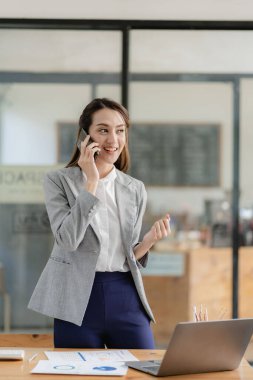 Gülümseyen Asyalı iş kadını masasının önünde akıllı telefonuyla iş tartışıyor.