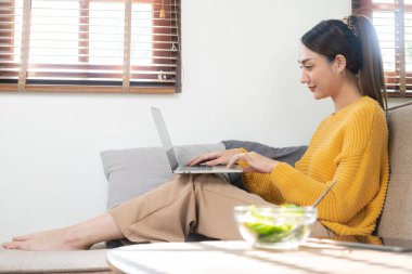 Hırslı Asyalı bir kadın kanepesinde oturmuş tablet bilgisayar ekranında çalışıyor gülümseyen bir kadın postayı kontrol ediyor ya da evde iletişim kurarken interneti arıyor.