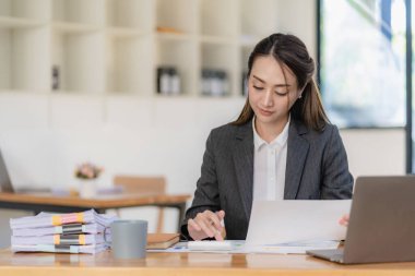 Parlak modern ofisindeki tablet bilgisayarında çalışan gülümseyen Asyalı iş kadını grafik belgeleriyle finansal rapor harcamalarını hesaplıyor.