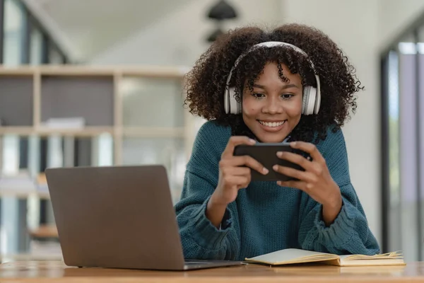 Female student in headphones holding smartphone, playing games and using mobile apps, online distance learning, watching video courses or zoom calls, making notes in workbook sitting at home.