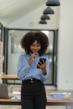Gülümseyen Afro-Amerikan kadın muhasebeci bilgisayarını kullanarak telefonda konuşuyor internet teknolojisinde oturuyor. Ofis iş konseptinde mobil iletişim