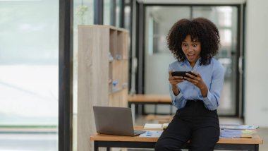 Gülümseyen Afro-Amerikan kadın muhasebeci bilgisayarını kullanarak telefonda konuşuyor internet teknolojisinde oturuyor. Ofis iş konseptinde mobil iletişim