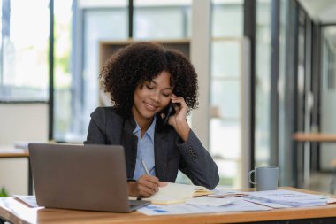 Gülümseyen Afro-Amerikan kadın muhasebeci bilgisayarını kullanarak telefonda konuşuyor internet teknolojisinde oturuyor. Ofis iş konseptinde mobil iletişim