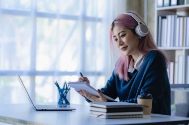 Asyalı, kulaklık takan bir kız öğrenci dizüstü bilgisayarla internetten ders çalışıyor internette çalışıyor, evde mutlu bir şekilde gülümsüyor..