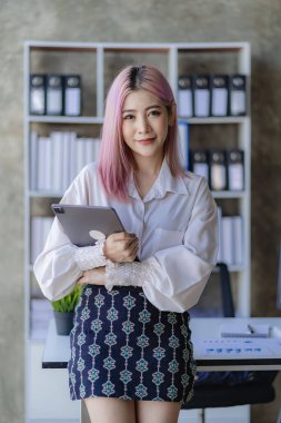 Çekici pembe saçlı Asyalı kadın çalışan masanın önünde duruyor. Dokunmatik tablet bilgisayar kullanıyor, gülümsüyor, iş bilgilerini şirketin ofisindeki bir masada müşterilere e-posta atıyor..