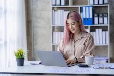 Finans ofisinde çalışan genç Asyalı muhasebeci hesap makinesi ve finansal belgeler kullanarak satış raporlarını yönetiyor..
