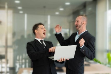 İki genç iş adamı dizüstü bilgisayara bakıp gülümsüyor ve iş planlarına göre başarılı olduğu için sevinçlerini dile getiriyorlar. Meslektaşları birbirleriyle iyi bir ortaklık hakkında konuşuyorlardı.