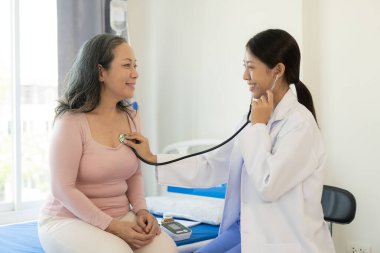 Asyalı yaşlı kadın mutlulukla gülümsüyor ve masada yaşlı bir kadınla konuşan bir kadın doktor tarafından teselli ediliyor. Muayenenin, muayenenin ve tedavinin sonuçlarını anlatıyor.