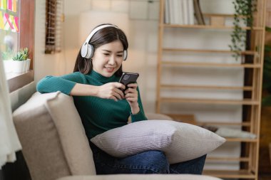Parlak bir dairede koltukta uzanmış, kulaklık takan Asyalı kadın evde dinlendirici müzik dinliyor..