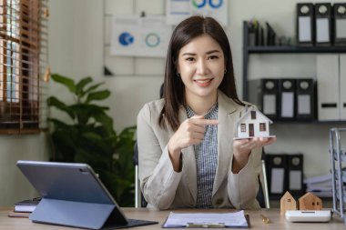 Çekici Asyalı kadın emlakçı ya da ev sigortacısı elinde bozuk para kavanozu ve tasarruf konseptiyle mini ev modelini tutuyor.