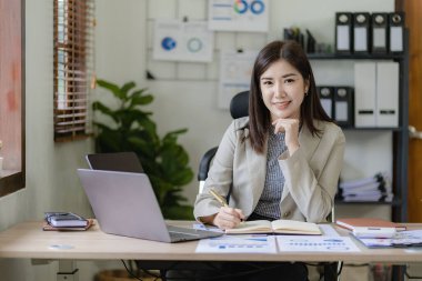 Masa, rapor, muhasebe, istatistik ve analitik araştırma kavramında finansal matematik yapmak için dizüstü bilgisayar ve hesap makinesi finansman grafikleri kullanan Asyalı güzel bir iş kadını.