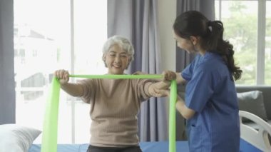 Genç bayan fizyoterapist, kas egzersizi ve fizyoterapi için direnç bantları kullanan gri saçlı yaşlı kadına kol hareketlerini gösteriyor.