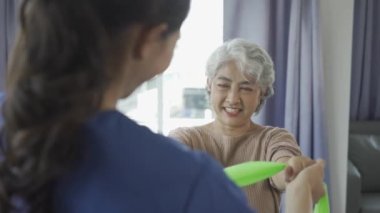 Genç bayan fizyoterapist, kas egzersizi ve fizyoterapi için direnç bantları kullanan gri saçlı yaşlı kadına kol hareketlerini gösteriyor.