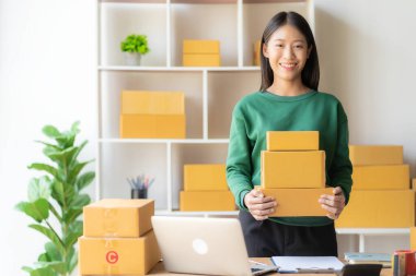 Heyecanlı bir Asyalı bayan küçük işletme sahibi sipariş almak ve paket teslim etmek için dizüstü bilgisayarını kullanırken odasında çalışıyor. KME iş fikri