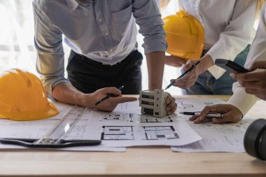 Mühendis takımı toplantısı ve konuşma, dizüstü planlar ve kağıtlar kullanılarak masada sarı şapkalı ev modeliyle birlikte, proje yöneticisi iş arkadaşıyla görüşüyor.