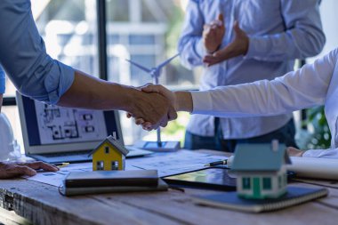 İki başarılı mühendis ya da mimar el ele tutuşurken güneş panelleri ve ev elektriği için rüzgar türbinleriyle masaya oturuyorlar..