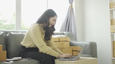Asyalı iş kadını çevrimiçi satış yapıyor ve dizüstü bilgisayardan, telefondan sipariş alıyor ve nakliye paketleriyle ilgili notlar alıyor. İçişleri Bakanlığı kadın girişimcisi (KOBİ))