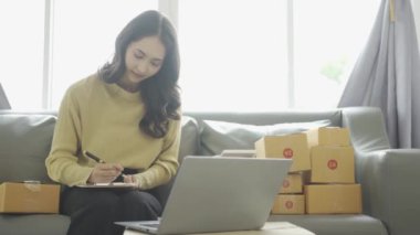 Asyalı iş kadını çevrimiçi satış yapıyor ve dizüstü bilgisayardan, telefondan sipariş alıyor ve nakliye paketleriyle ilgili notlar alıyor. İçişleri Bakanlığı kadın girişimcisi (KOBİ))