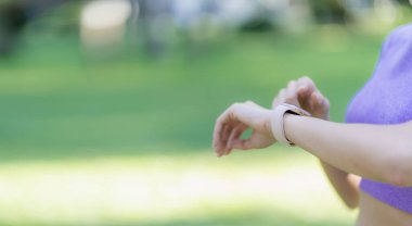 Asyalı kadın parkta ya da bahçesinde yeşil bitkilerle egzersiz yaparken ayakta durup saatine bakıyor, spor konsepti