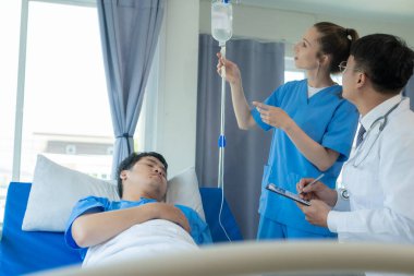 Bir kadın doktor ve iki erkek doktor, genç bir erkek hastayı yatakta tedavi ediyorlar ve hastane yatağının yanında, acil serviste Asyalı bir erkek hastayla konuşuyorlar..