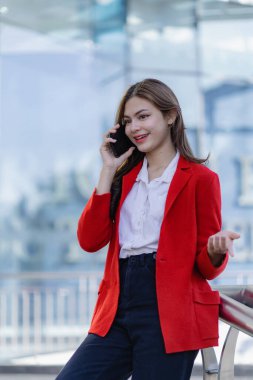 Çekici bir kadın mutlu mesut ayakta duruyor ve ofis binasının yanındaki telefonda iş hakkında konuşuyor. Gülümseyen kadın elinde akıllı telefonla dışarıda yürüyen müşteriyle konuşuyor.