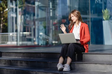 Bilgisayarı elinde tutan kendine güvenen bir kadın kameraya bakıyor. Gülümseyen dişler şirketin yanında duruyor. Açık havada dizüstü bilgisayarla çalışıyor.