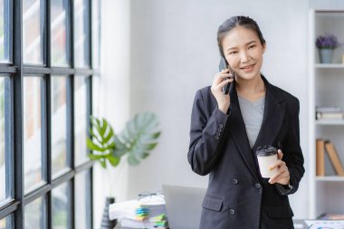 Çekici Asyalı iş kadını ofisteki iş ortağıyla telefonda konuşuyor. Cep telefonu ve dizüstü bilgisayarlı mali grafik belgeleri müşterilerin iş adamlarının konseptiyle iletişime geçmesini bekliyor.