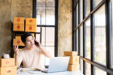 Akıllı telefon ve dizüstü bilgisayarla online satış yapan Asyalı genç işadamı çevrimiçi müşterilerden sipariş alıyor, paket teslimatlı KOBİ iş kavramı.