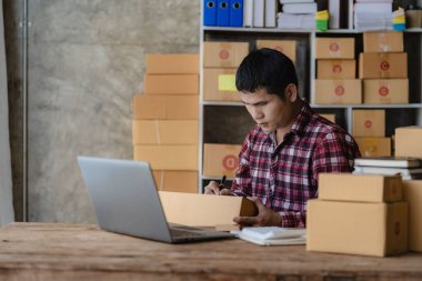 Depoda çalışan genç Asyalı iş adamı, merkez ofis, paket kutularıyla bağımsız kurye şirketi, müşteri adreslerini dizüstü bilgisayarla kontrol ediyor..