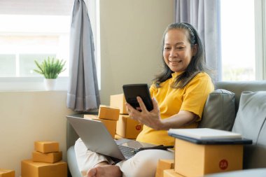 Küçük bir işletme KOBİ kurun. Akıllı telefon ya da laptop tableti kullanan olgun yaşlı kadın elleri kutuları hazırlamak için çevrimiçi siparişleri alır ve onaylar. çevrimiçi alışveriş kavramı