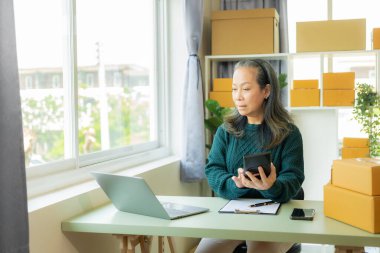 Küçük bir işletme KOBİ kurun. Akıllı telefon ya da laptop tableti kullanan olgun yaşlı kadın elleri kutuları hazırlamak için çevrimiçi siparişleri alır ve onaylar. çevrimiçi alışveriş kavramı