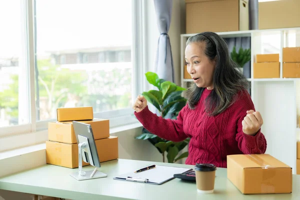 Successful old businesswoman smiling happily receiving new buzzer Asian woman owns a small business online with shipping boxes. concept of working at home