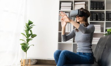 VR kulaklıklı genç Latin kadının havaya dokunmasına şaşırdım. Gözlüklü gülümseyen beyaz kadın sanal gerçeklik teknolojisi aygıtı koltukta otururken 3D simülasyon videosu izliyor..
