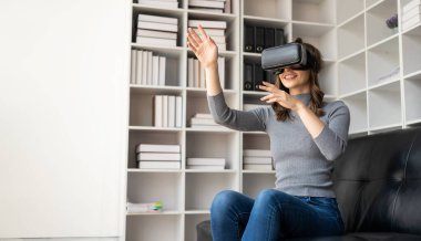 VR kulaklıklı genç Latin kadının havaya dokunmasına şaşırdım. Gözlüklü gülümseyen beyaz kadın sanal gerçeklik teknolojisi aygıtı koltukta otururken 3D simülasyon videosu izliyor..
