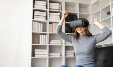 VR kulaklıklı genç Latin kadının havaya dokunmasına şaşırdım. Gözlüklü gülümseyen beyaz kadın sanal gerçeklik teknolojisi aygıtı koltukta otururken 3D simülasyon videosu izliyor..