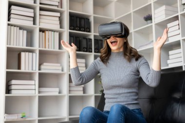VR kulaklıklı genç Latin kadının havaya dokunmasına şaşırdım. Gözlüklü gülümseyen beyaz kadın sanal gerçeklik teknolojisi aygıtı koltukta otururken 3D simülasyon videosu izliyor..