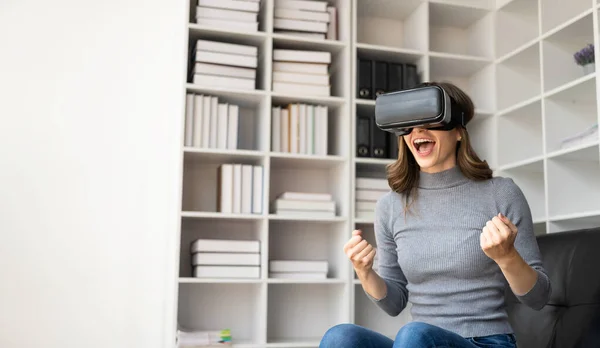 stock image Surprised young latin woman in VR headset touching the air. Smiling caucasian woman wearing vr glasses virtual reality technology device watching 3d simulation video while sitting on sofa.