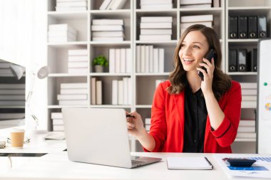 Kırmızı tişörtlü genç bayan dizüstü bilgisayarla çalışıyor ve akıllı telefondan konuşuyor ortağıyla bir görüşme ayarlamak için iş görüşmesi yapıyor. Teknoloji kullanma kavramı