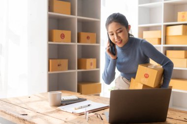 Küçük Girişim, KOBİ, Girişimci - Akıllı telefon veya dizüstü bilgisayar kullanan mutlu Asyalı kadın, kutu hazırlamak ve çevrimiçi siparişleri iletmek için çevrimiçi siparişleri alır ve onaylar.