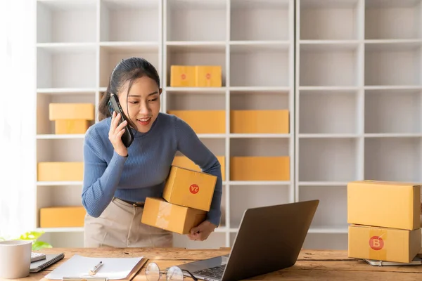 Small Business Startup, SME, Entrepreneur - Happy Asian woman using smartphone or laptop to receive and verify online orders to prepare boxes and deliver parcels from online order.