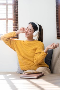 Sarı tişörtlü, mutlu, kulaklık takan ve hafta sonu evde olan oturma odasında müzik dinlemenin keyfini çıkaran güzel bir Asyalı kız..