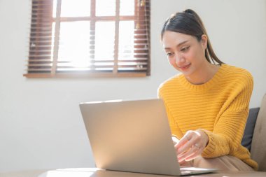 Sarı tişörtlü güzel Asyalı kız evde dizüstü bilgisayarla çalışıyor. Sosyal medya pazarlamasında çalışan serbest çalışan kadın, küçük işletme girişimi fikri.