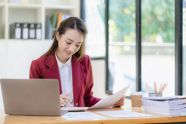 stock image Asian businesswoman in suit sitting on desk in office, with computer document graph for bookkeeping in workplace to calculate annual profit by function, business concept