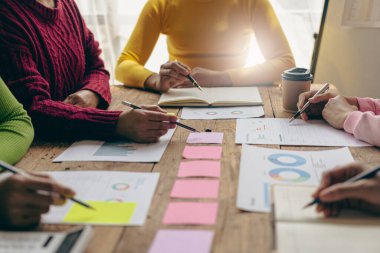 Bir grup Asyalı genç iş adamı, günlük kıyafetleriyle finansal veri grafiklerini analiz ediyor ve iş toplantısında beyin fırtınası yapıyorlar.