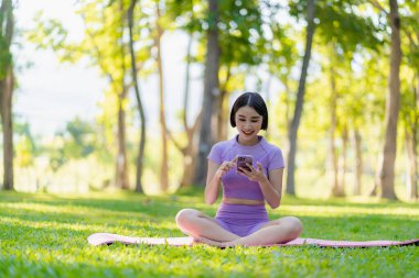 Asyalı bir kadın akıllı telefonuna bakıyor, parkta yoga yapıyor sağlıklı genç bir kadın yoga yapıyor. Vücut, akıl ve sağlık için doğal çevrede yoga yapıyor. Rahatla konsept.
