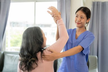 Asyalı yaşlı kadın hemşire desteğiyle fizyoterapi yapıyor. Genç kadın huzurevinde omuz kasları için fizyoterapi yapıyor.