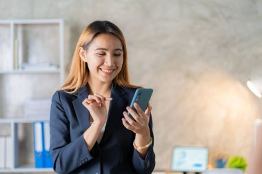 Asyalı iş kadını iş yerindeki müşterileriyle konuşurken akıllı telefon tutuyor. Hesap makinesi, iş bilgisayarı, mali muhasebeci, kredi bankası, hesap makinesi ya da doküman kullanıyor.,