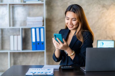 Asyalı iş kadını iş yerindeki müşterileriyle konuşurken akıllı telefon tutuyor. Hesap makinesi, iş bilgisayarı, mali muhasebeci, kredi bankası, hesap makinesi ya da doküman kullanıyor.,