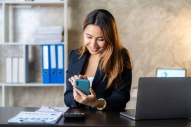 Asyalı iş kadını iş yerindeki müşterileriyle konuşurken akıllı telefon tutuyor. Hesap makinesi, iş bilgisayarı, mali muhasebeci, kredi bankası, hesap makinesi ya da doküman kullanıyor.,